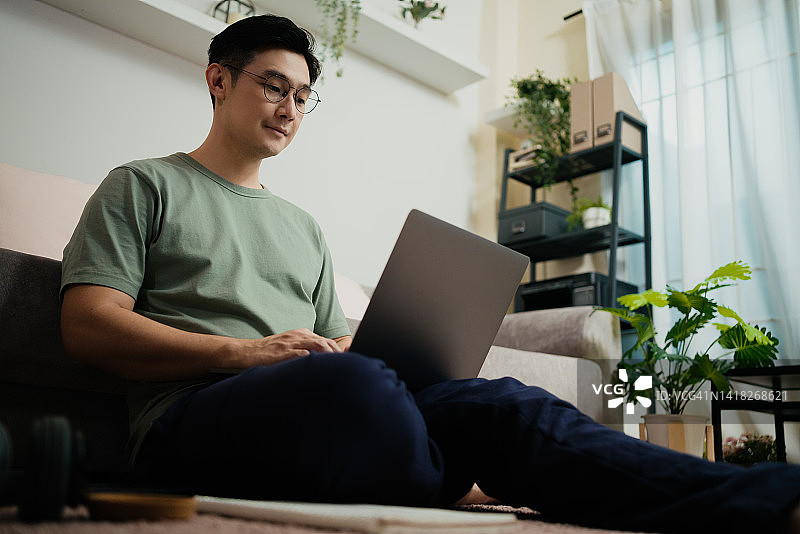亚洲成年学生在家中客厅的沙发上用笔记本电脑在线学习并喝热咖啡图片素材
