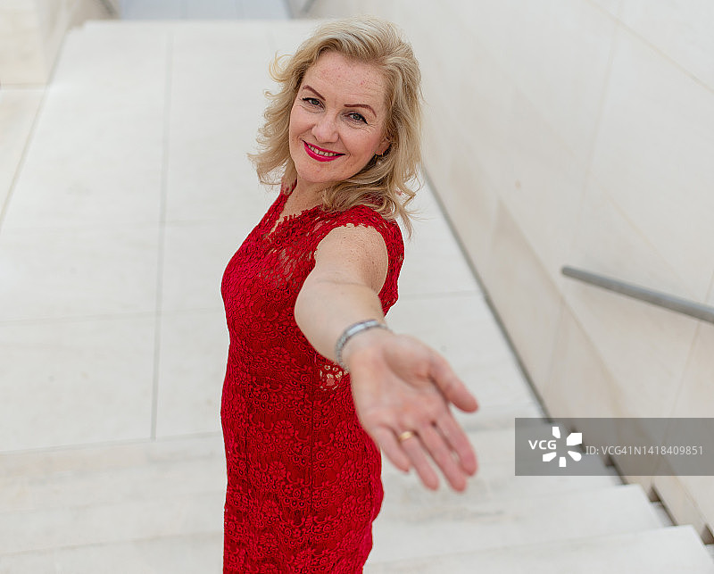 一位身穿红色连衣裙的女士走在楼梯上，并用手邀请您的侧面图。有魅力的中年妇女穿着连衣裙在楼梯上用手邀请您与她同行图片素材