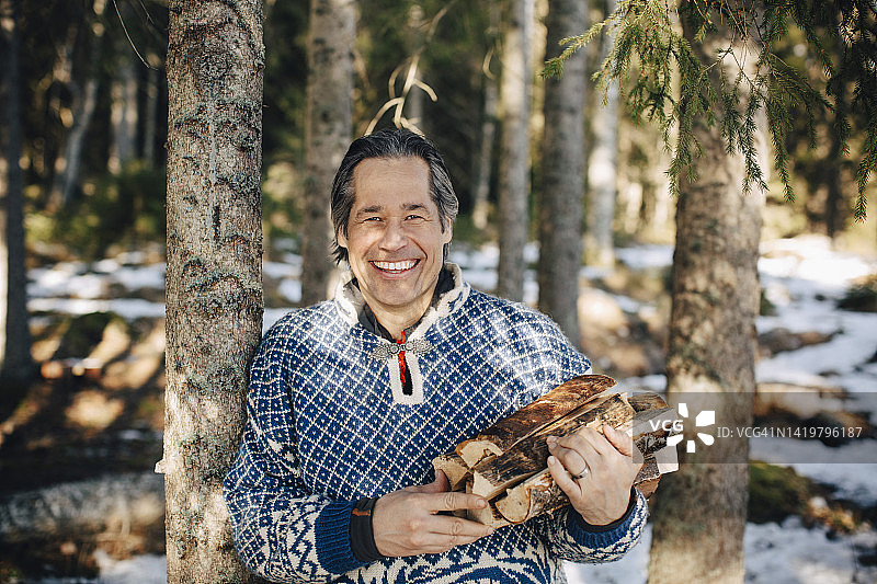 快乐的成熟男子拿着木柴站在森林里的树旁的肖像图片素材