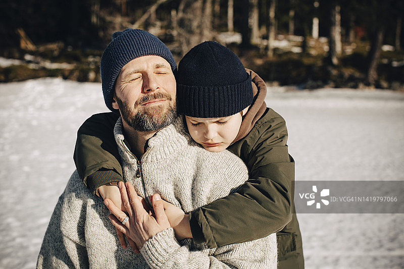 冬日阳光下，男孩闭着眼睛拥抱穿着暖和的父亲图片素材