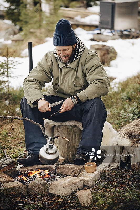 在篝火旁看着茶壶的穿暖和衣服的微笑男子图片素材