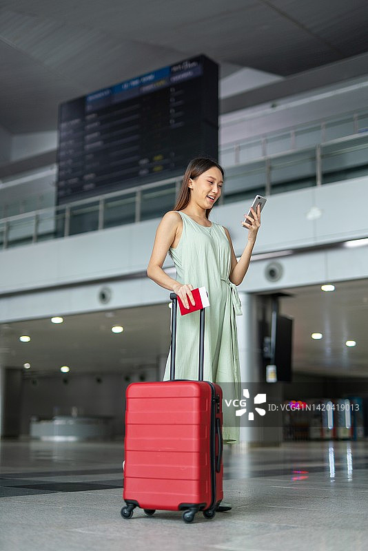 年轻亚洲女性手持手机和行李箱走过出发指示牌，前往梦想的度假目的地图片素材