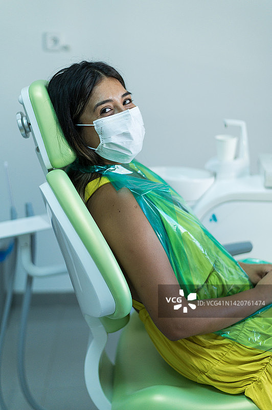 戴着口罩坐在牙科诊所，看着镜头的病人肖像。坐在牙科椅上的女病人侧面照图片素材