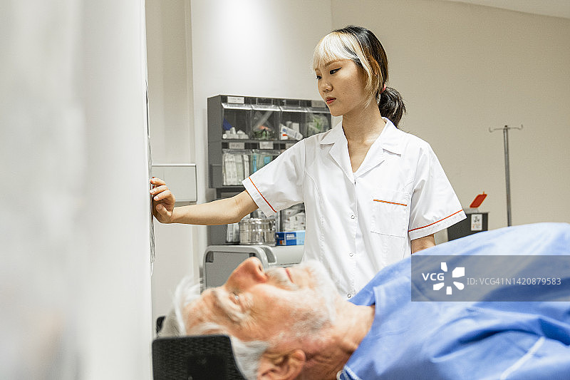 老年男性患者正在接受核磁共振扫描，女性操作员在背景中的侧面视角图片素材