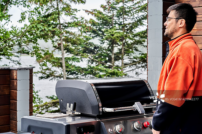 穿着橙色卫衣站在烤架旁欣赏森林和湖景的亚洲男人图片素材