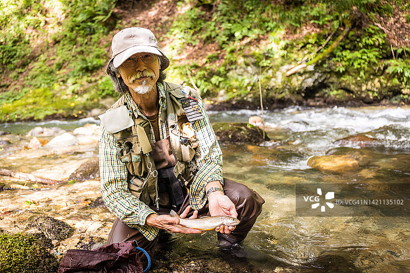 在山间河里飞蝇钓鱼的老年男人展示他捕获的鱼（Iwana）图片素材