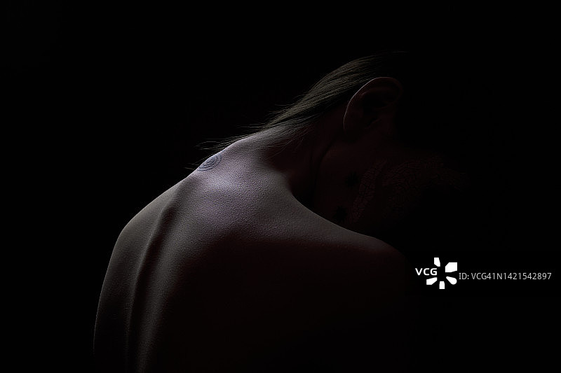 从后面看，一位有纹身的亚洲年轻女子向前倾着头。脊椎骨强烈突出，在黑色孤立的背景上形成美丽的图案。现代艺术、颈椎骨软骨病、颈部和背痛的概念。图片素材
