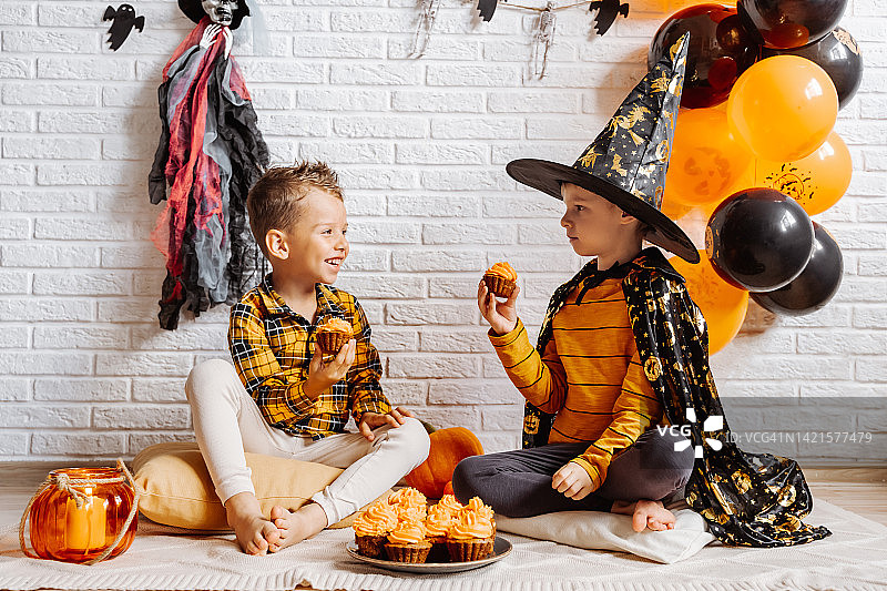 两个小男孩拿着纸杯蛋糕庆祝万圣节，背景是装饰过的白色砖墙图片素材