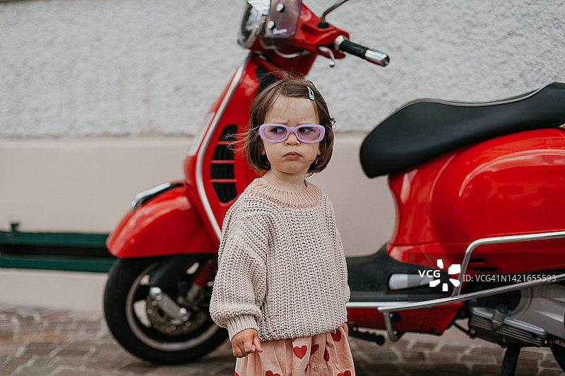 一个穿着时髦衣服的蹒跚学步的小女孩的肖像，旁边是一辆红色老式摩托车图片素材