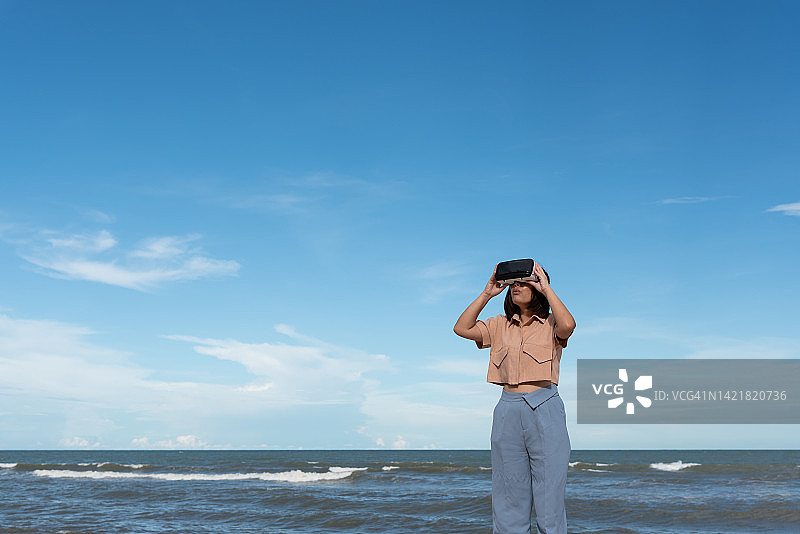 站在海滩上，使用VR设备看天空的年轻亚洲女性图片素材