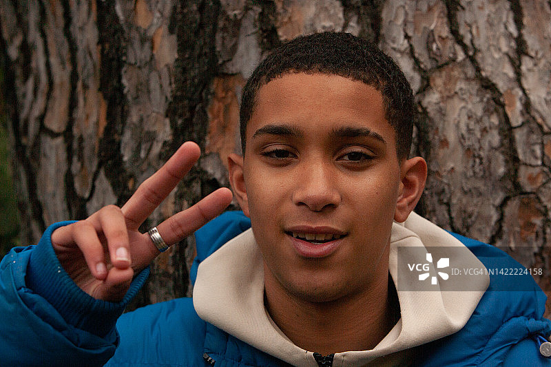 一位英俊的年轻人用他的手做出和平的标志，一个穿着蓝色和白色休闲装、留着短款非洲发型的十几岁男孩对着镜头微笑，同时用他的手和手指做出胜利的V字标志图片素材