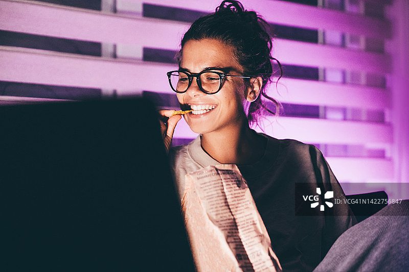 夜晚女人用笔记本电脑看电影吃玉米片图片素材