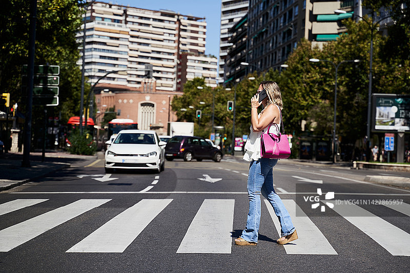中年白人妇女在城市人行横道上边过马路边用智能手机交谈。技术与生活方式概念图片素材