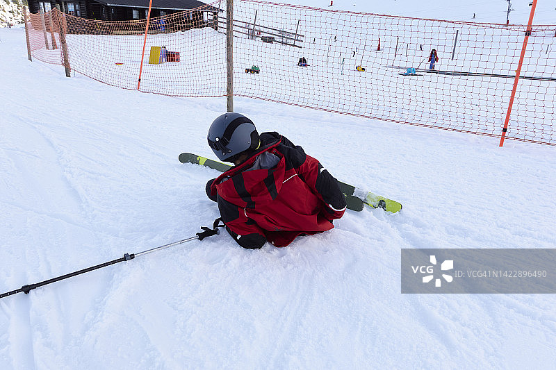滑雪者摔倒在雪地，后方可见防护网和木屋图片素材