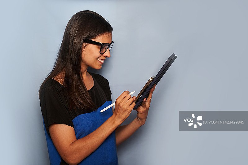 戴眼镜的女人在浅蓝色背景下拿着平板电脑微笑地看着屏幕。带有复制空间的轮廓视图。图片素材