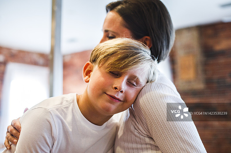 男孩享受母亲的爱，头靠在她的肩膀上，闭着眼睛，微笑着图片素材
