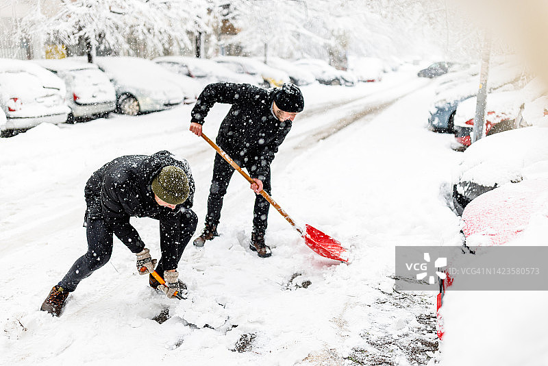 两个年轻人在车前用铲子扫雪。图片素材