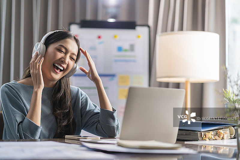 快乐的亚洲休闲商务女性在办公桌前工作时听着耳机里的音乐，玩得很开心。放松的年轻女性戴着耳机，使用笔记本电脑播放音乐播客，微笑的女办公室职员戴着耳机图片素材