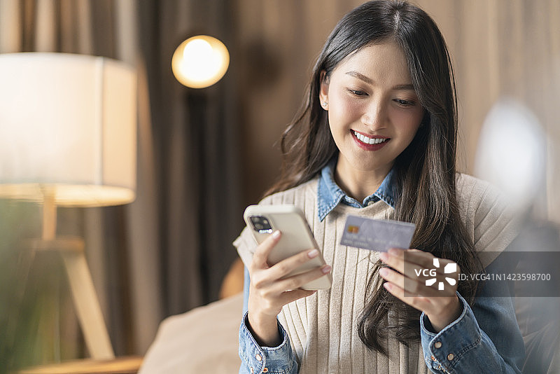 快乐的亚洲年轻成年女性在家里客厅的沙发上用信用卡通过智能手机在线购物，享受购买折扣服装的乐趣图片素材