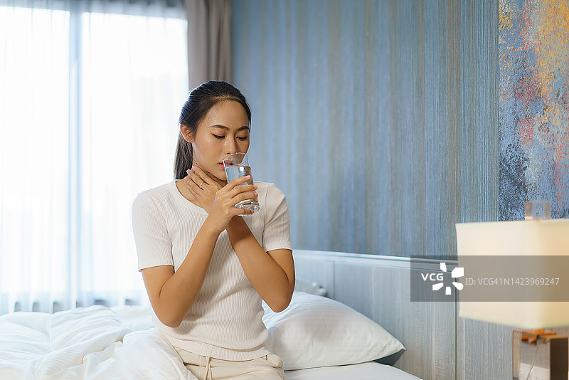 喉咙痛的亚洲女人穿着睡衣在卧室的床上喝水缓解图片素材