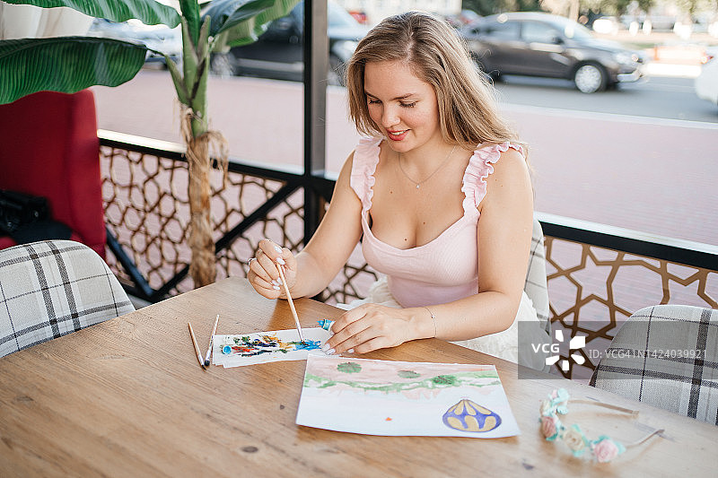 在夏日咖啡馆里用颜料和画笔在纸上绘画的美丽女孩图片素材