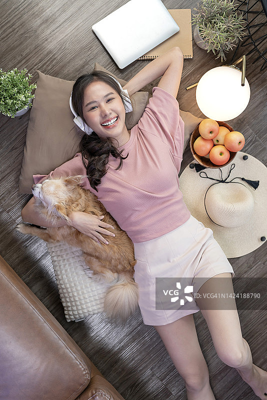 年轻女子周末居家休闲放松，戴着耳机躺在木地板上，与吉娃娃宠物狗为伴图片素材