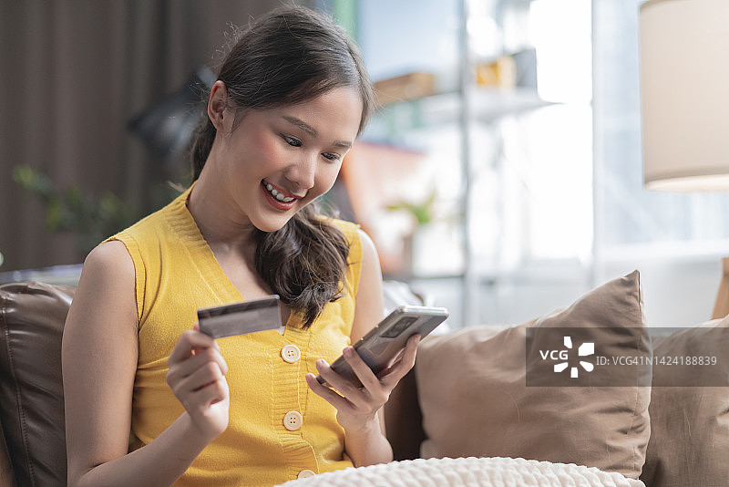 快乐的亚洲年轻成年女性在家里客厅的沙发上用信用卡通过智能手机在线购物，享受购买折扣服装的乐趣图片素材