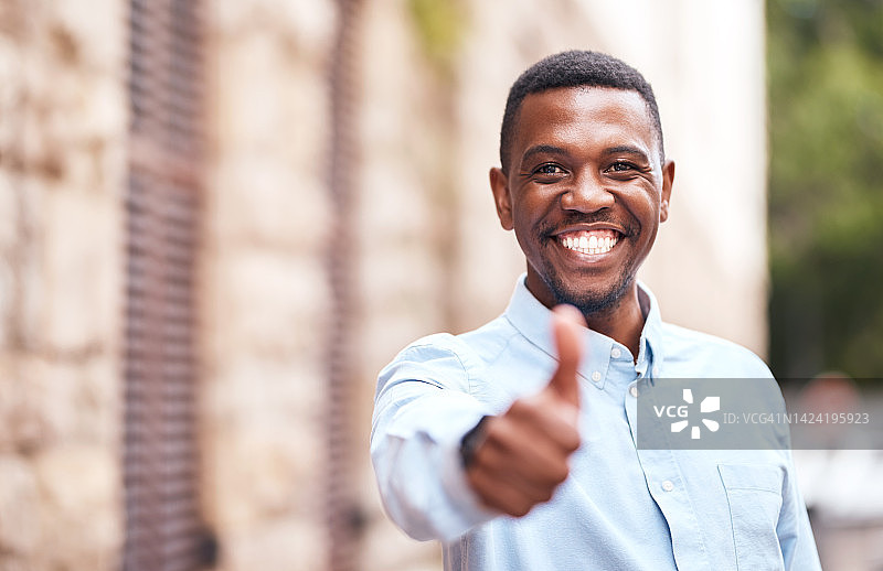 在户外城市街道上微笑并竖起大拇指的非洲男子肖像。在建筑物外做出点赞或同意手势的快乐、兴奋和积极的黑人男子图片素材