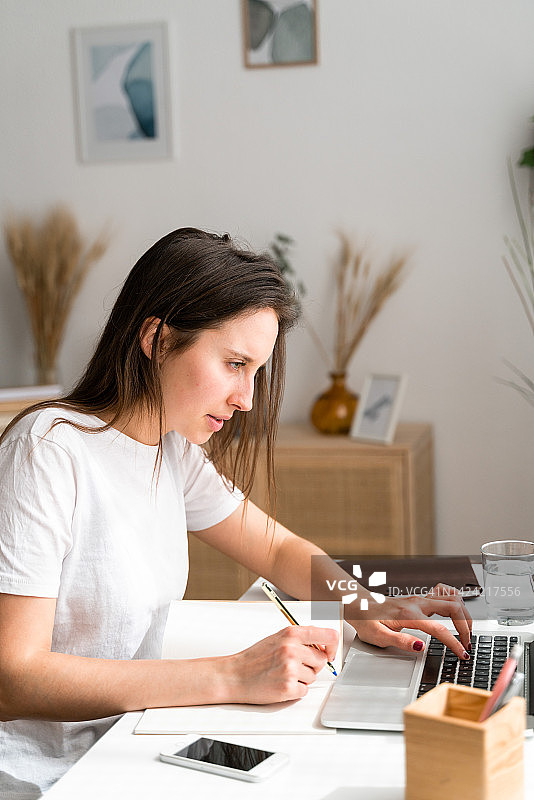 专注的女人在家使用笔记本电脑工作并做笔记图片素材