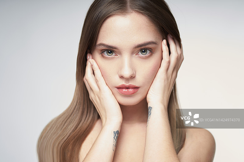 年轻美女肖像：浅灰色背景，飘逸长发，肌肤良好，手臂有纹身。无衬衫金发女郎手靠近脸庞，裸妆和自然美，皮肤护理，头发护理。图片素材