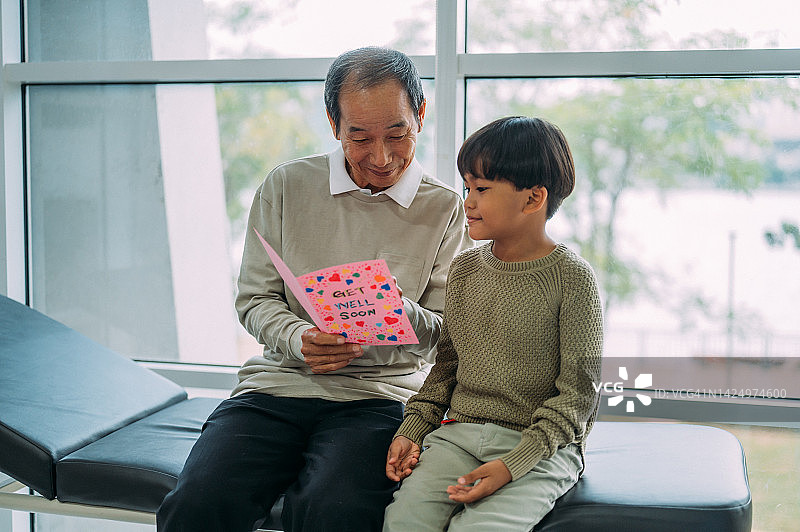 年长的亚洲男病人在医院病房里和孙子一起阅读慰问卡图片素材