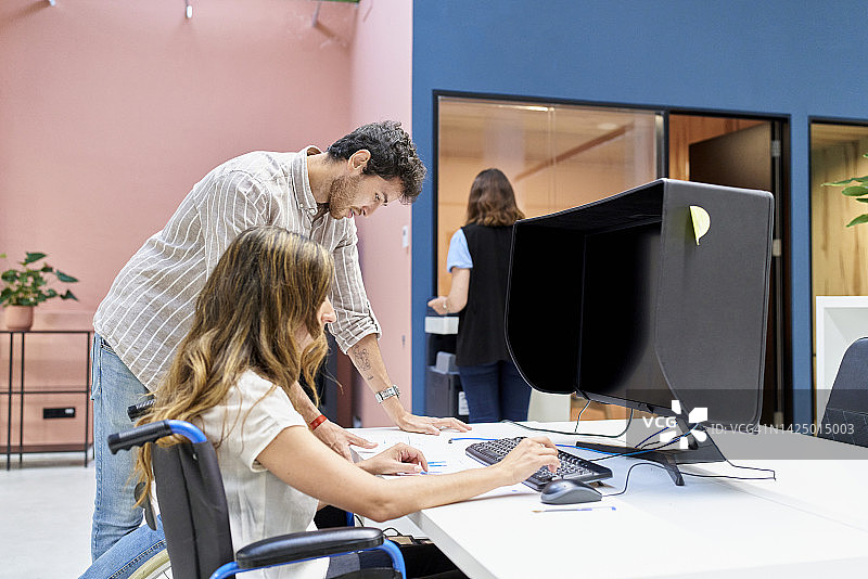 年轻女人坐在轮椅上，在电脑前工作，同事站立审阅文件，另有一女子站在其身后图片素材