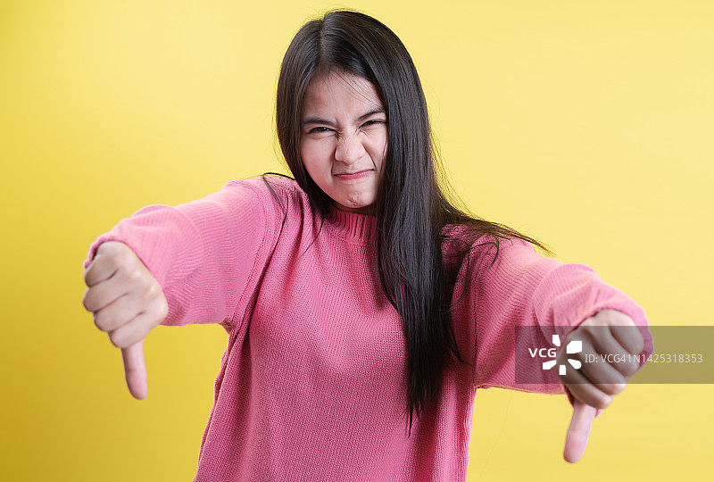 不满的亚洲女人表示不赞同，拇指向下，表达不喜欢，不满地皱着眉头，在黄色背景下隔离。肢体语言概念。图片素材