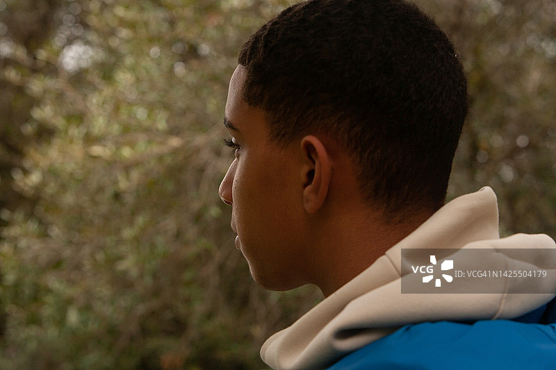 英俊的年轻男子在自然环境中，略微从后方看到他的侧面，一个穿着蓝色羽绒服和奶油色连帽衫的青少年，从他的有利位置在天然绿色植物中若有所思地向外看图片素材