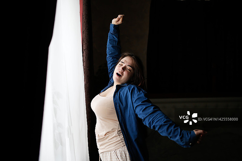 超重的美丽女人在早晨的窗户边伸展身体，慵懒的早晨，黑暗的背景图片素材