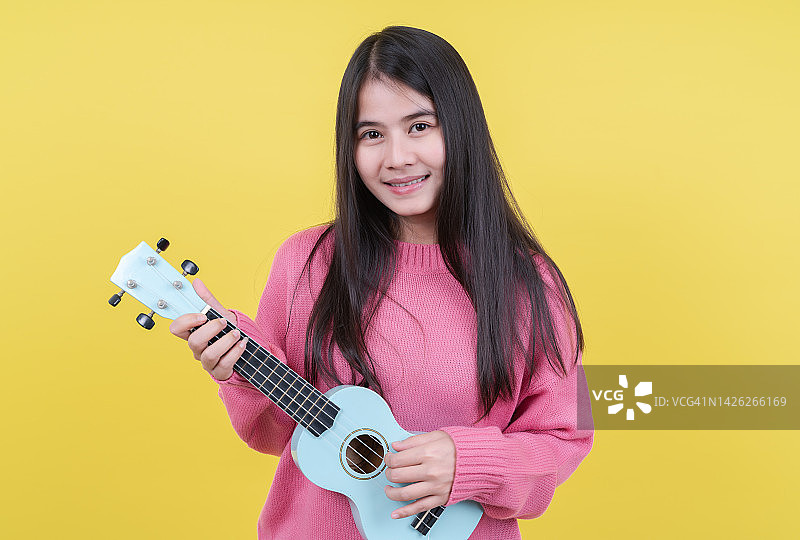 抱着尤克里里的快乐女孩站在粉色背景前，微笑的女音乐家弹奏尤克里里。图片素材