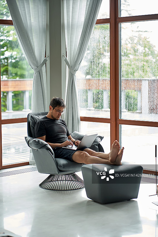 一位韩国男人坐在扶手椅上使用笔记本电脑，窗外下着雨图片素材