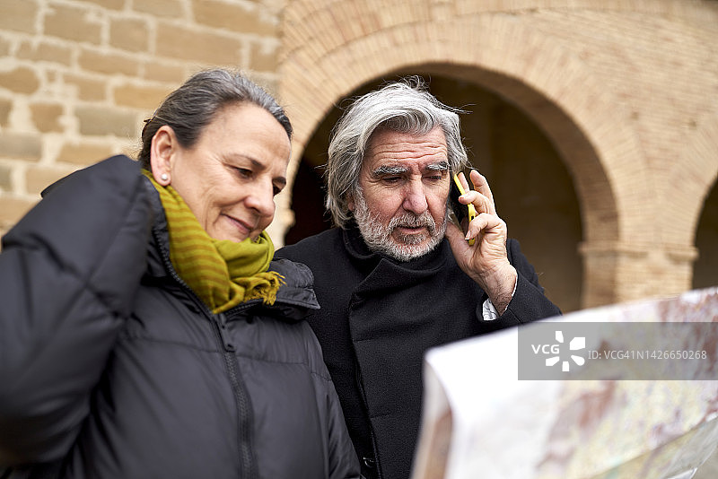 年长的男人在用手机交谈，他的妻子在看地图，寻找他们度假时将要访问的地方的地址图片素材