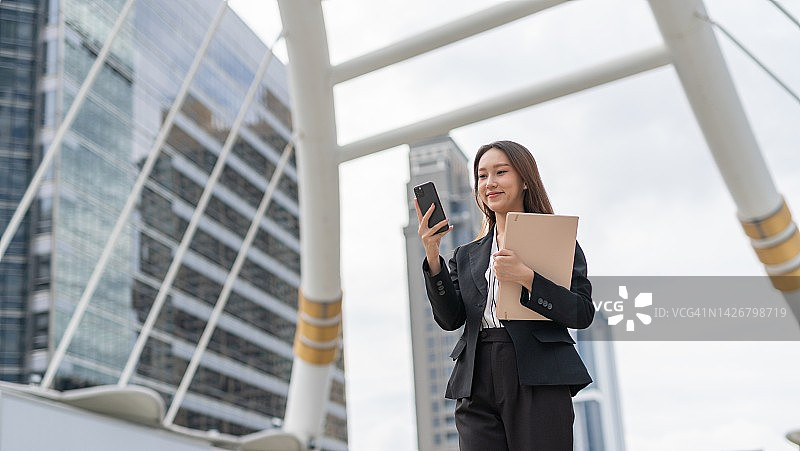 在曼谷中央商务区行走的，手持文件夹看手机的年轻亚洲女商人图片素材
