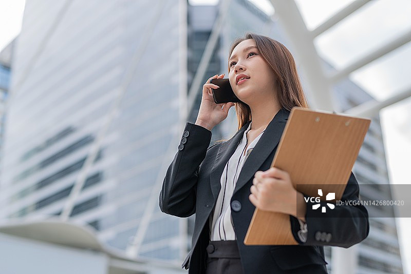 在曼谷中央商务区，拿着文件夹通过电话了解会议和销售库存报告最新情况的年轻有魅力的亚洲职业女商人图片素材