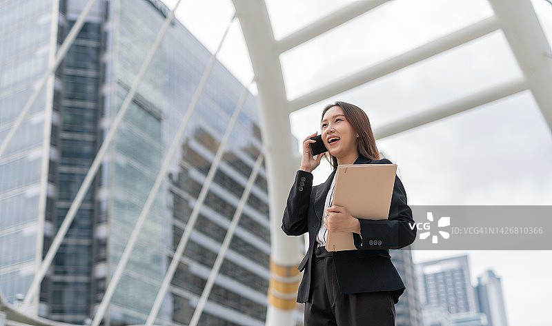 在曼谷中央商务区，笑容可掬的年轻亚洲女商人边走路边打电话，手里拿着文件夹图片素材