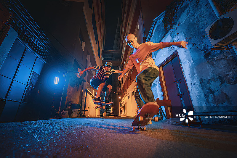 多元化的年轻男人夜间在小巷与朋友滑板腾空跳图片素材