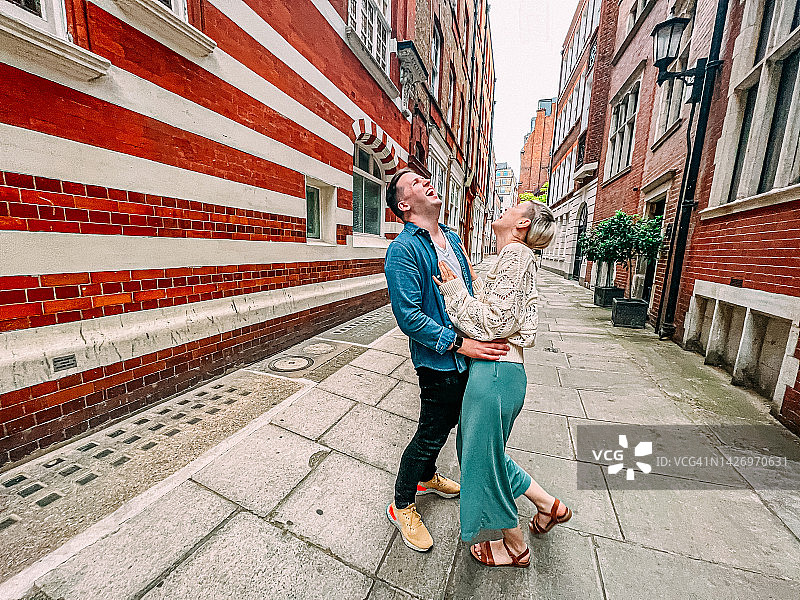 在英国伦敦色彩鲜艳的小巷中，一对可爱的时尚白人游客情侣在度假时站着欢笑的抓拍肖像图片素材