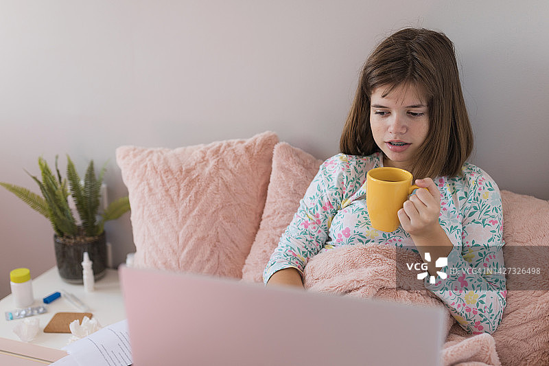 生病时一边用笔记本电脑上网一边喝茶的严肃女孩图片素材