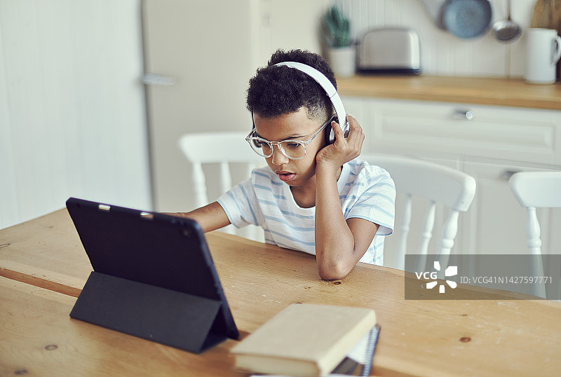 戴着无线耳机和眼镜的非洲小男孩在家中厨房的桌旁使用平板电脑图片素材