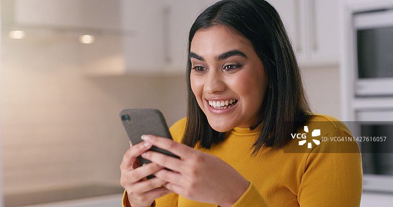 在厨房里用手机微笑的女人，在家里观看有趣的社交媒体文本或视频。女孩因好消息或网络交流而感到兴奋和快乐。图片素材