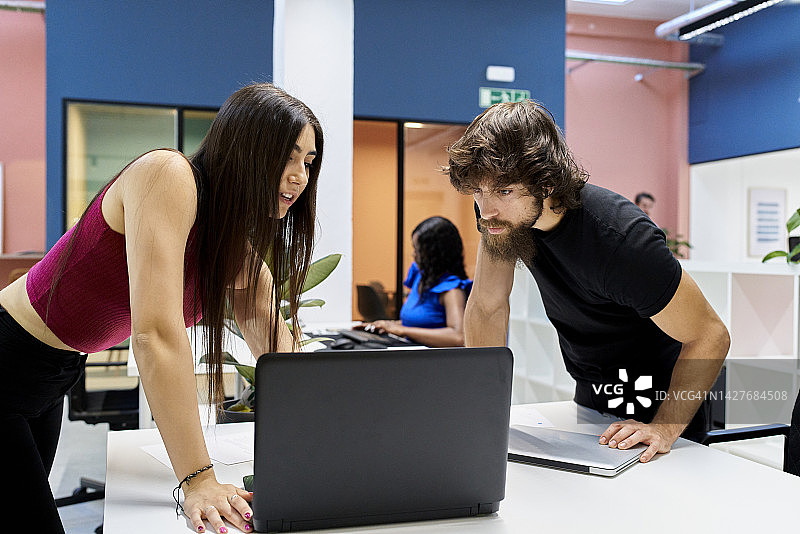 长发女子和胡须男子站在办公桌前使用电脑，身后有几个人在工作图片素材