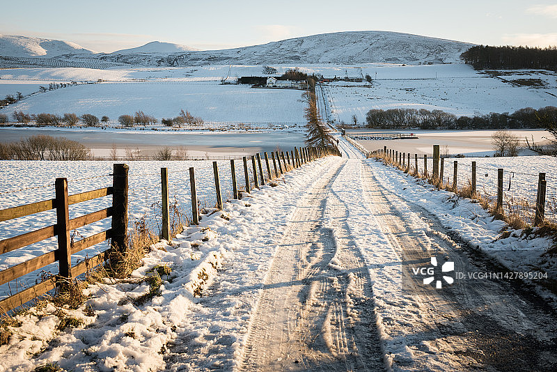 通往山丘的道路上，雪覆盖的木栅栏旁的轮胎痕迹，位于英国苏格兰爱丁堡的彭特兰山丘区域公园图片素材