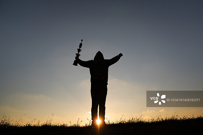 商人在岩石山峰上攀登，并在日落时举起奖杯庆祝胜利的剪影，成功、赢家、领导者概念图片素材