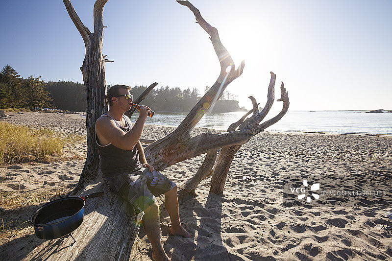 一名男子坐在海滩的圆木上喝啤酒图片素材